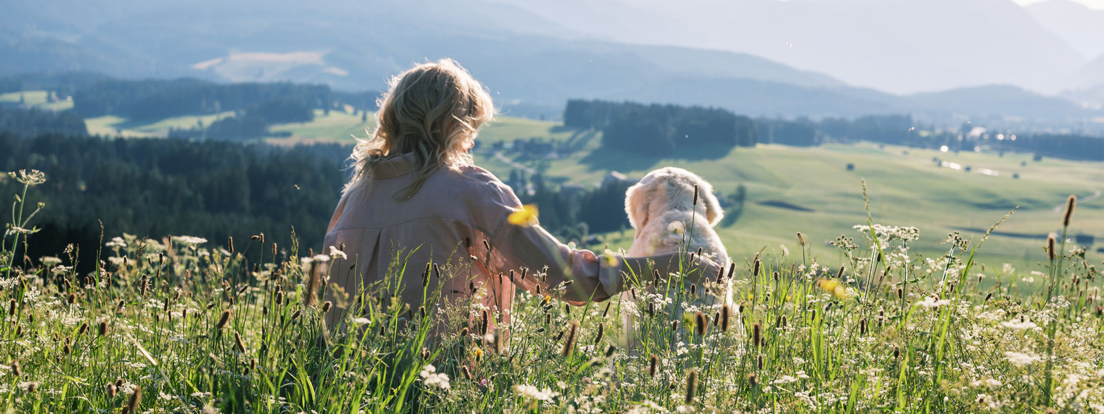 Katharina und Nala sitzen auf einer Wiese und schauen von einem Hügel auf die Wiese und Wälder die weiter unten liegen.